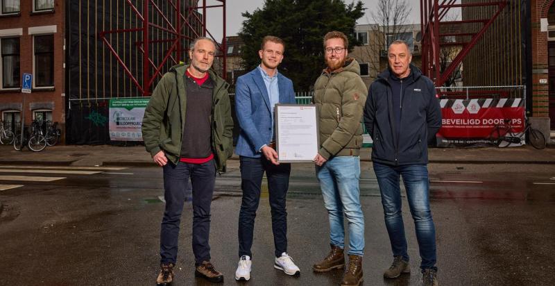 Vier mannen met op de achtergrond de bouwplaats aan de Claes de Vrieselaan. De twee mannen in het midden houden het certificaat vast.