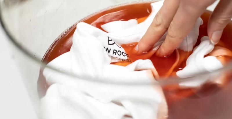 Op de foto een hand die een t-shirt in een kom verf duwt.