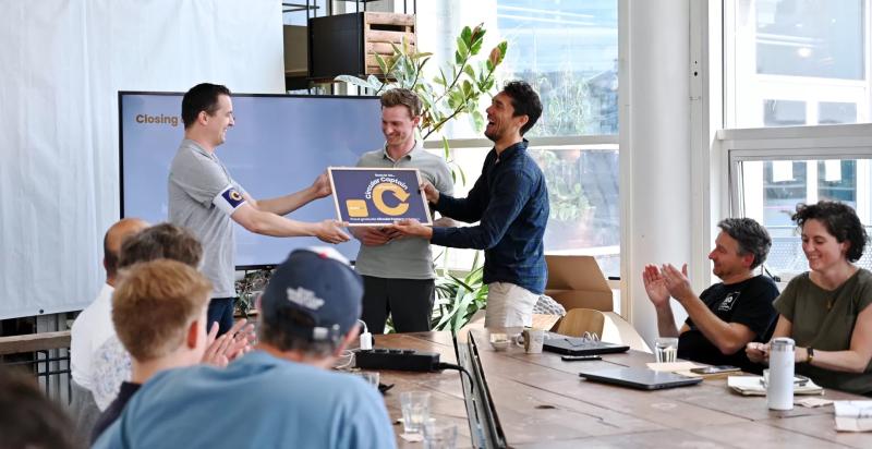 Op de foto drie mannen voor een tafel. De man links overhandigd een certificaat aan de man rechts.