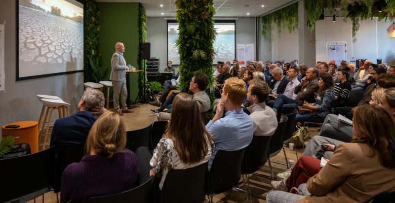 Grote groep mensen luistert in een grote zaal naar een spreker die een presentatie houdt.