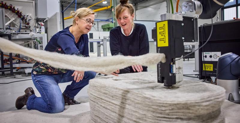 twee vrouwen zitten achter een machine die een lange witte streng wol vilt in de vorm van een stoel