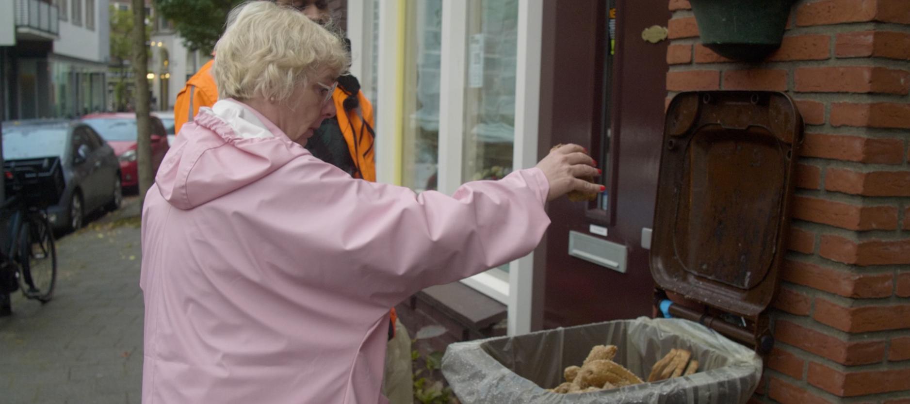 Wilma van der Wilt in een straat bij een bruine container, gooit brood in de container. Achter Wilma een gemeentemedewerker in oranje jas, kijkt toe.