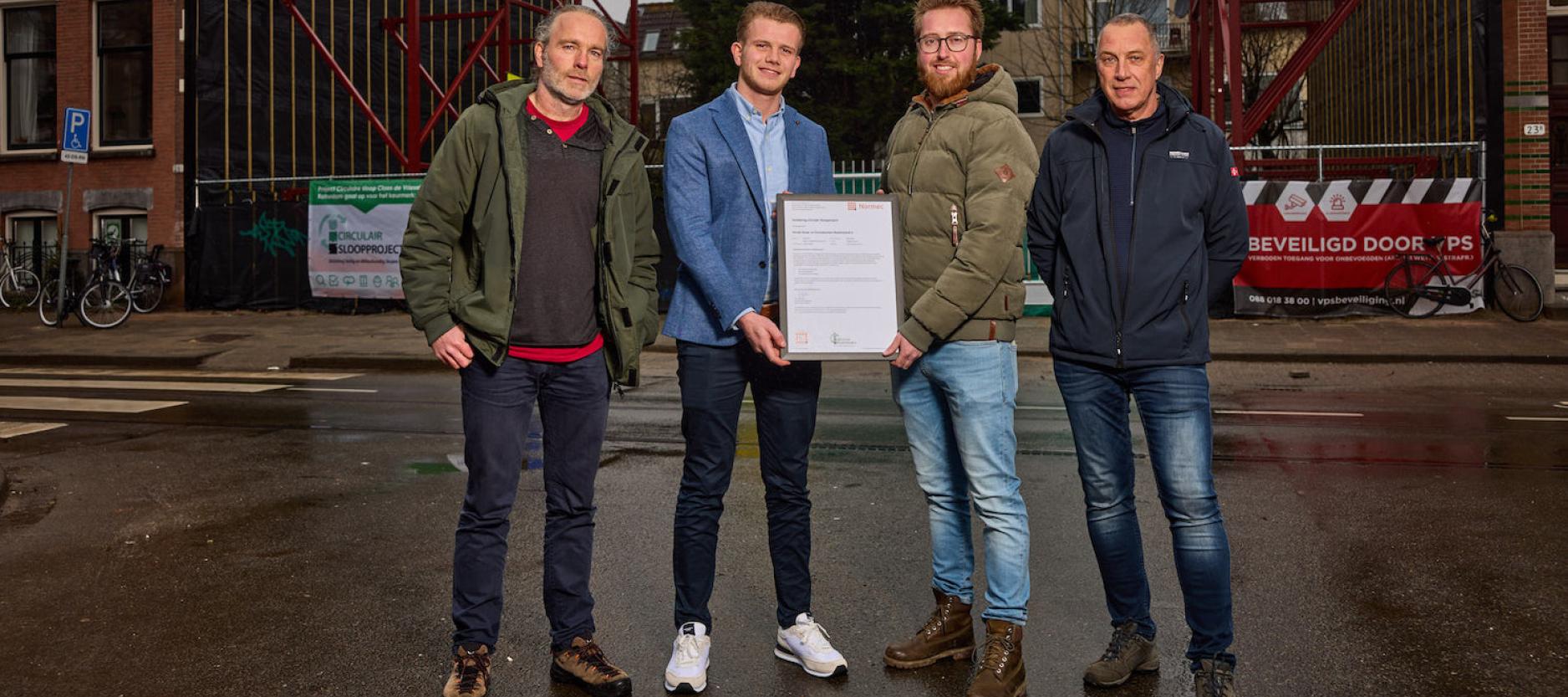 Vier mannen met op de achtergrond de bouwplaats aan de Claes de Vrieselaan. De twee mannen in het midden houden het certificaat vast.