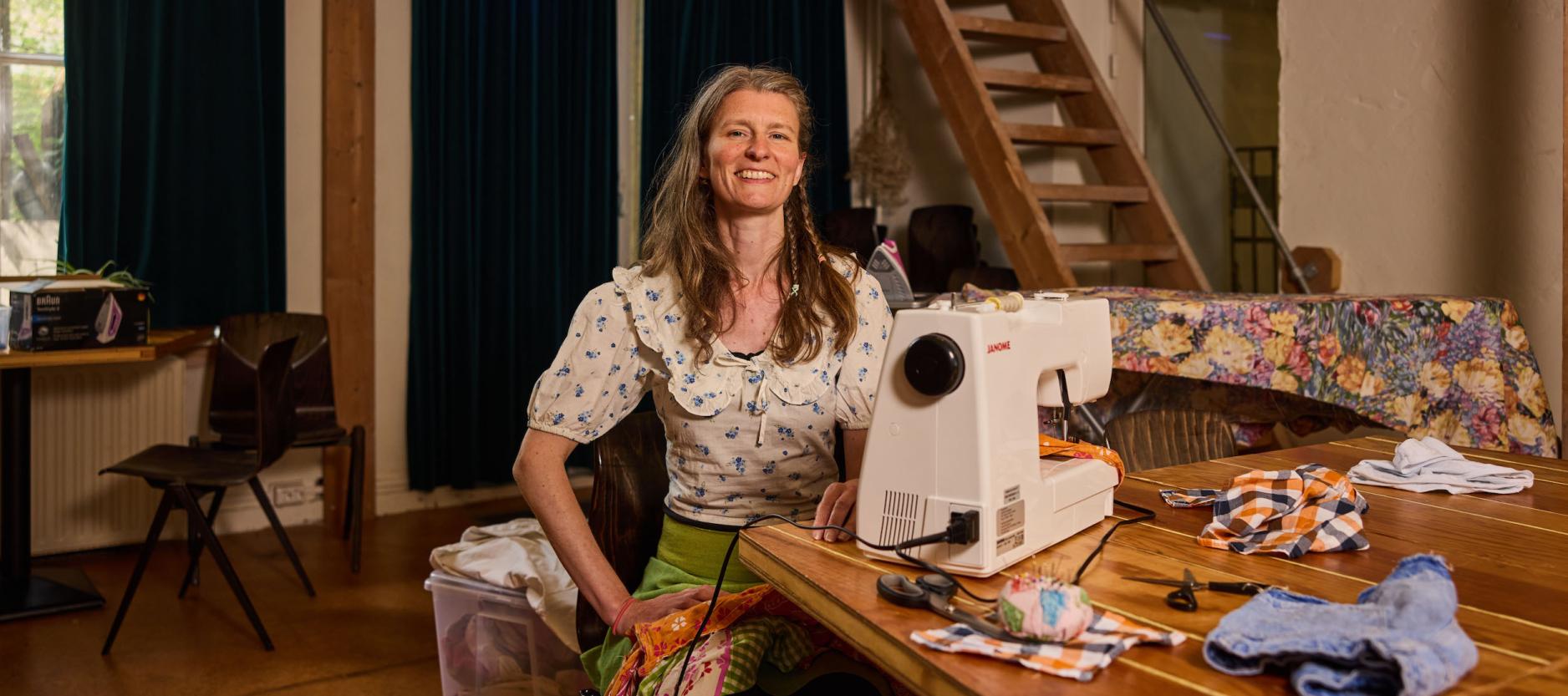 Kunstenaar Emma Hazenzak, poseert in huis aan een houten tafel in huis achter de naaimachine.