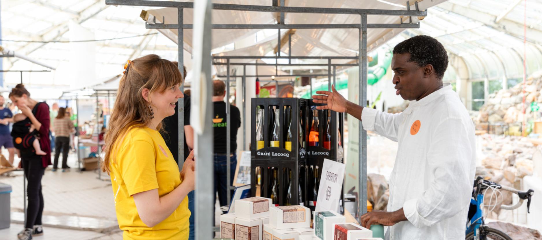 Man en vrouw staan bij een kraam, de man vertelt de vrouw over zijn wijnen