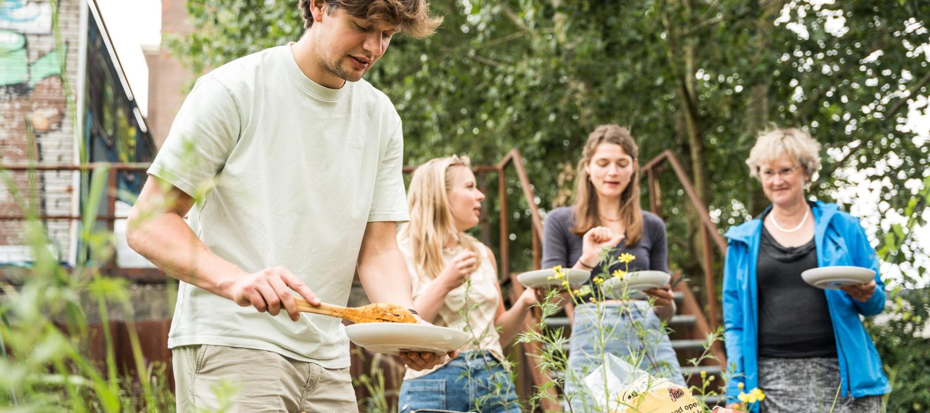 Mensen koken op een barbecue en hebben een bordje vast. Ze zijn in de natuur
