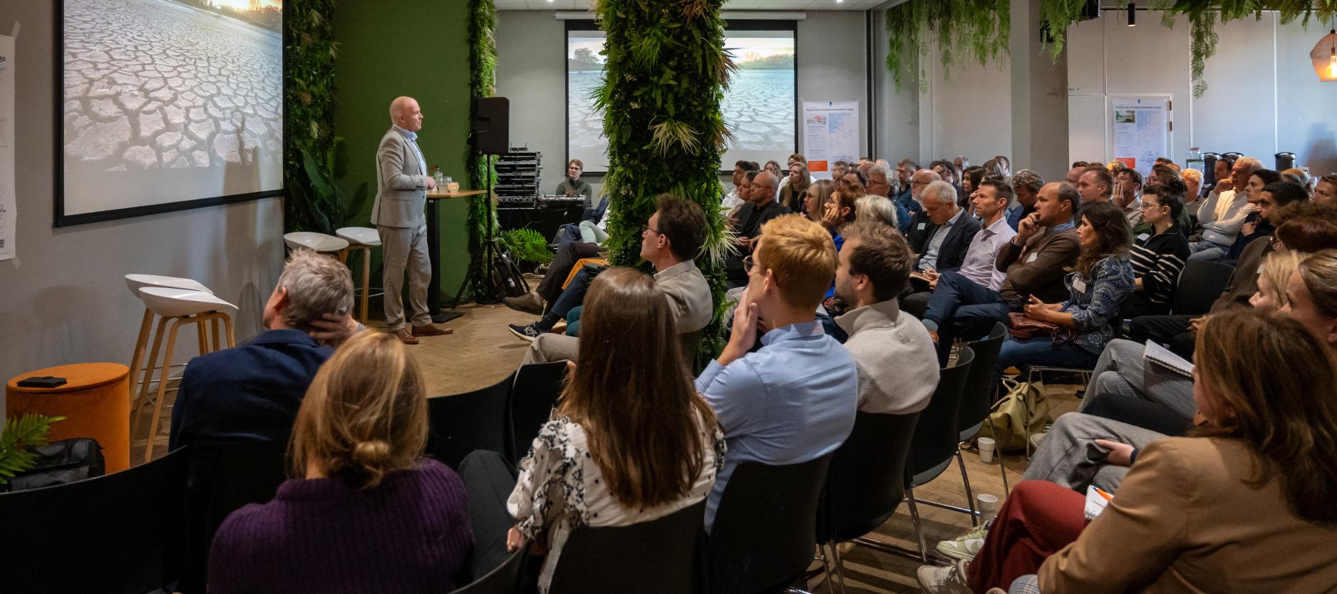 Grote groep mensen luistert in een grote zaal naar een spreker die een presentatie houdt.