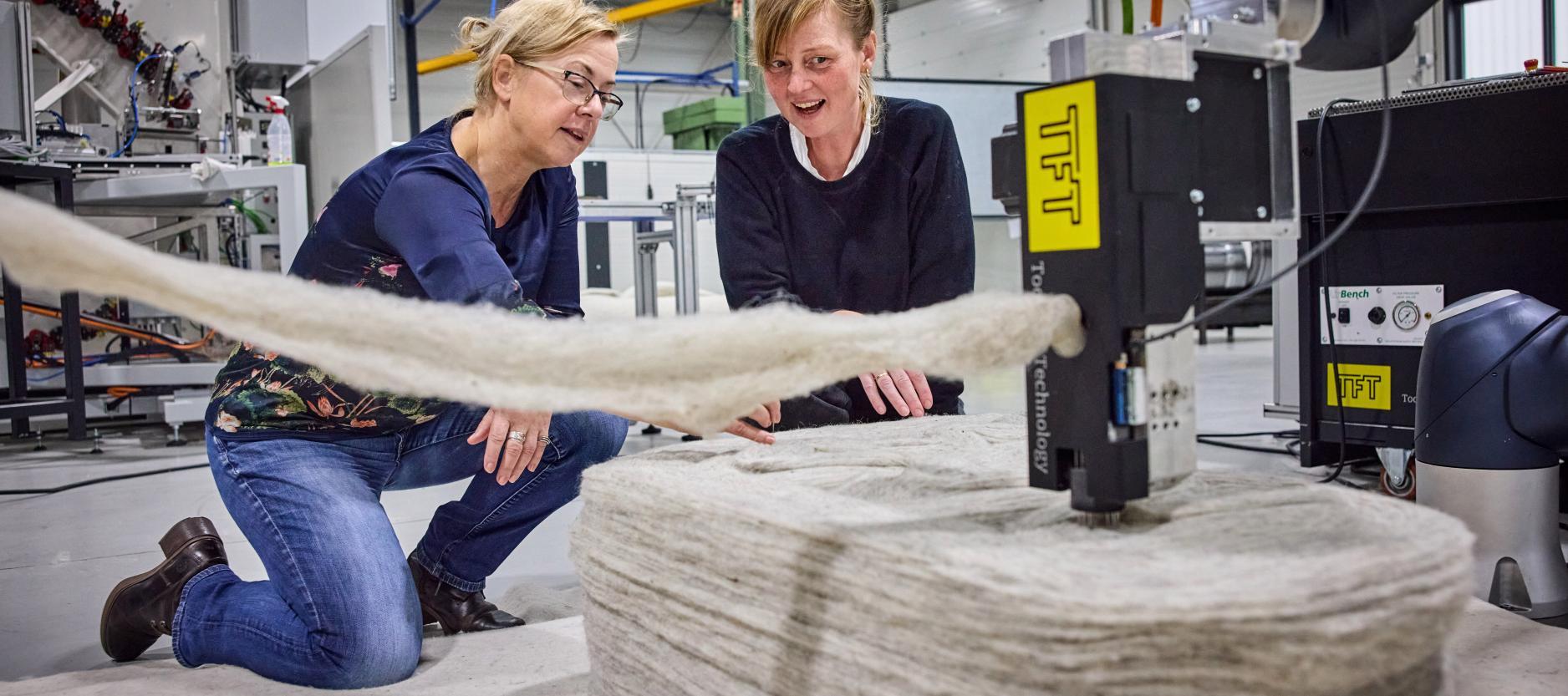 twee vrouwen zitten achter een machine die een lange witte streng wol vilt in de vorm van een stoel