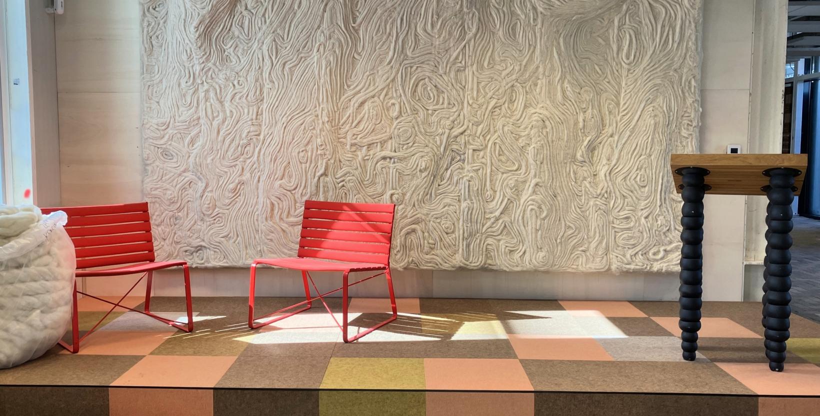 Podium met twee rode stoelen en aan de wand erachter een groot wollen wandpaneel.