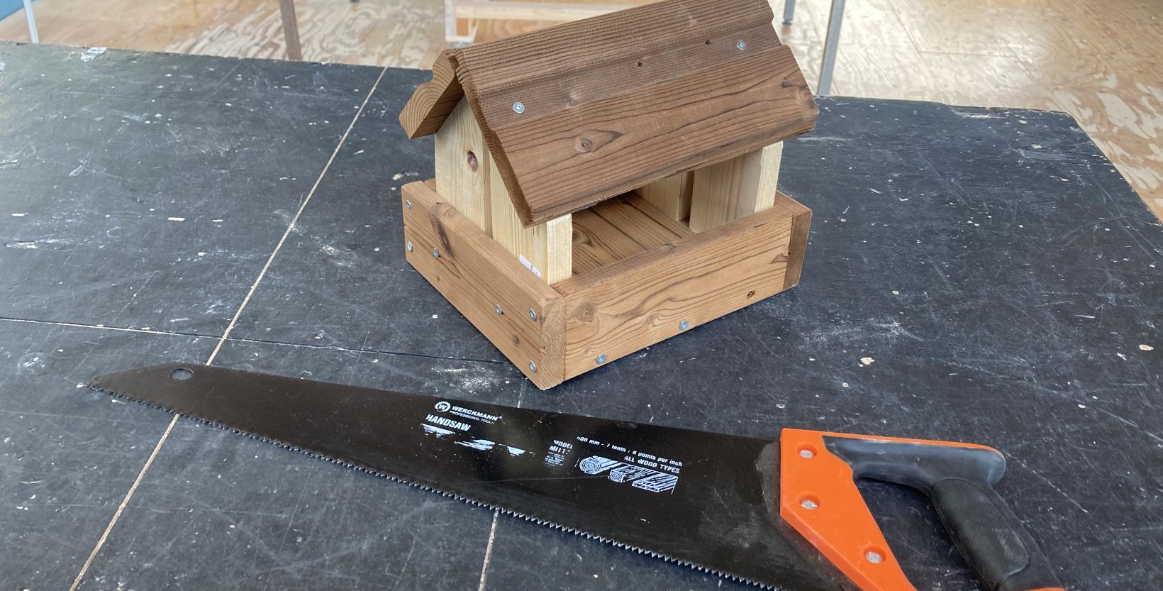 Tafel met een zaag erop en een houten voederhuisje voor vogels.