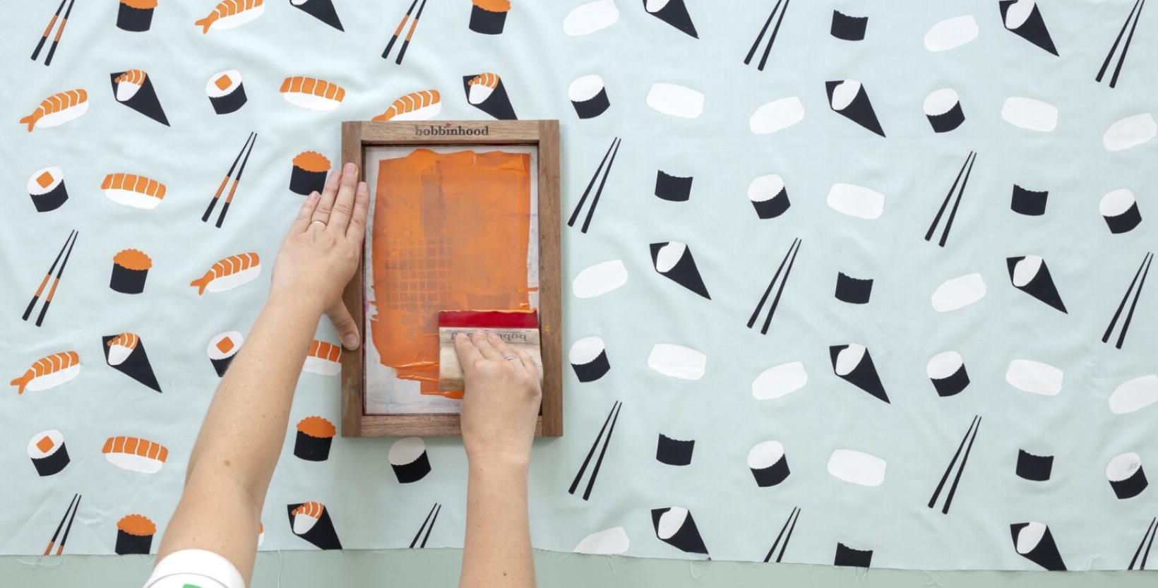 Bovenaanzicht van een tafel en twee handen die een zeefdruk aan het maken zijn.