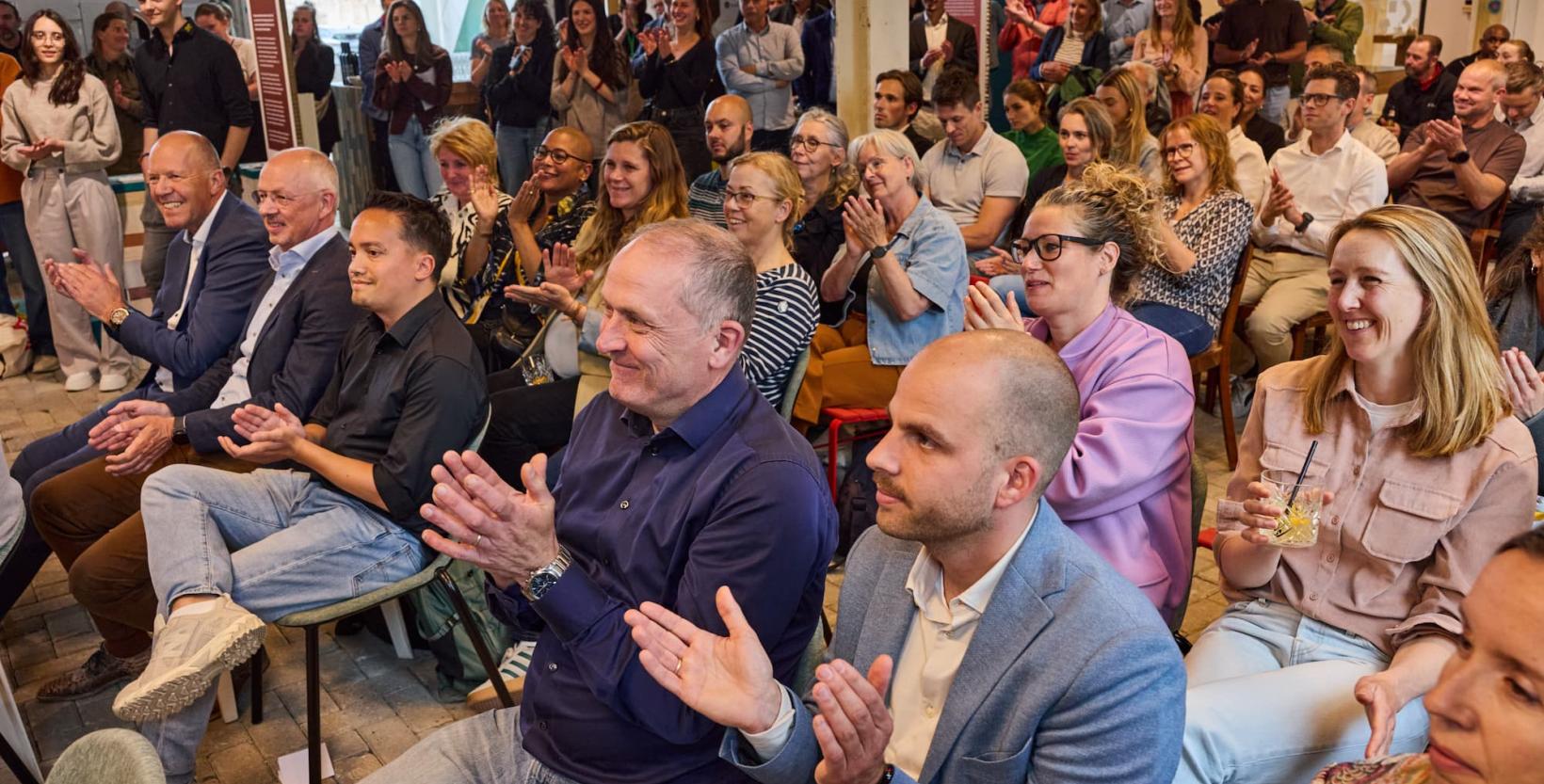 Een groep enthousiaste mensen geeft applaus tijdens een bijeenkomst in circulair centrum De HER. 