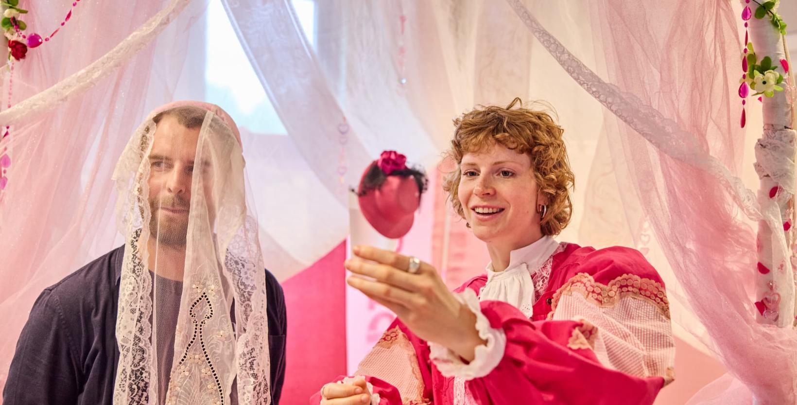 Een man met een sluier staat naast een zogenaamde trouwambtenaar met een roze outfit.