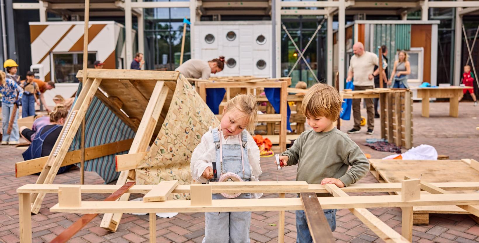 Twee kleine kinderen timmeren met hout. Op de achtergrond is een deel van de voorgevel van circulair centrum De HER te zien.