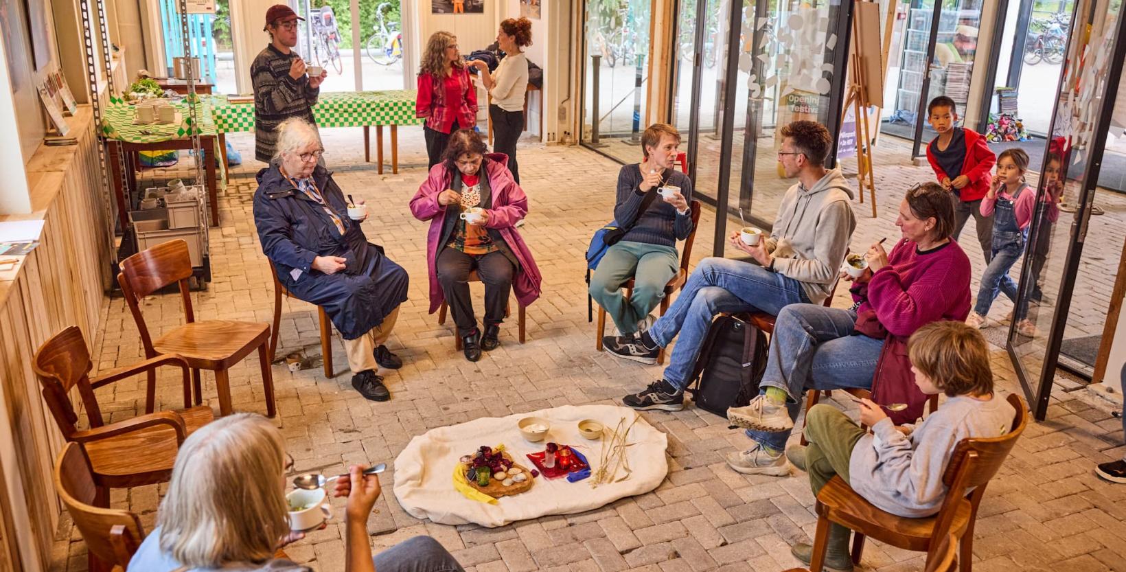 Een groep mensen zit op stoelen rond een kleedje met eten erop.