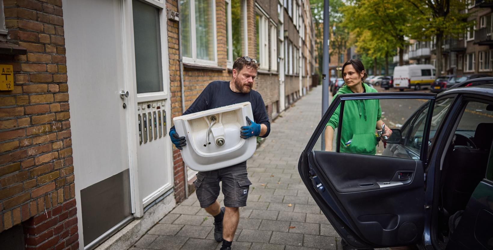 Man tilt een wastafel uit een woning, terwijl een vrouw naast een auto de deur openhoudt.