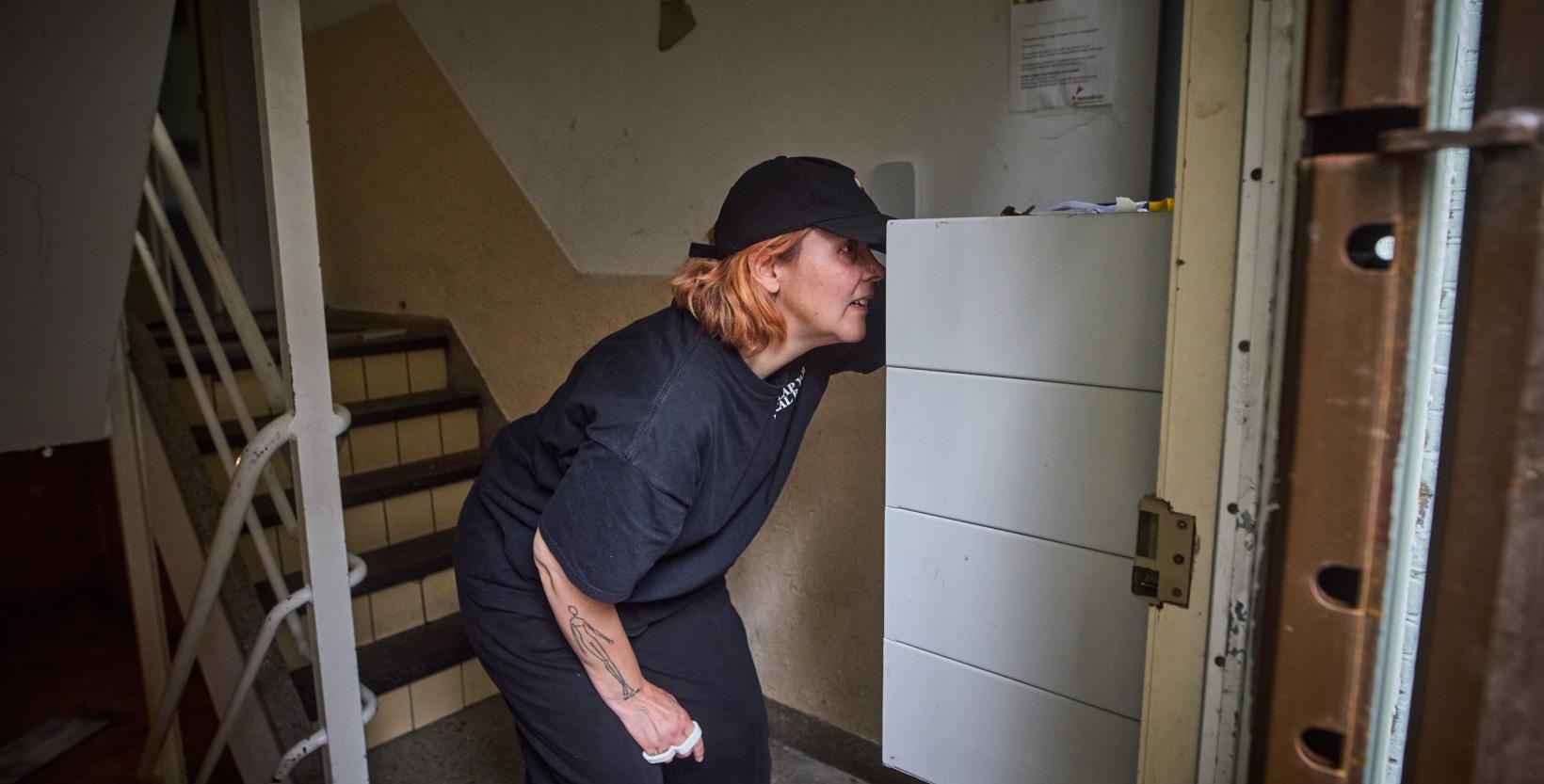 Vrouw in zwarte kleding en pet kijkt in de brievenbus in de hal.