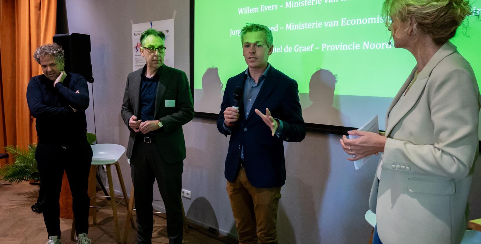 Drie heren staan voor een presentatiescherm op een congres en worden door de vrouwelijke dagvoorzitter bevraagd. 